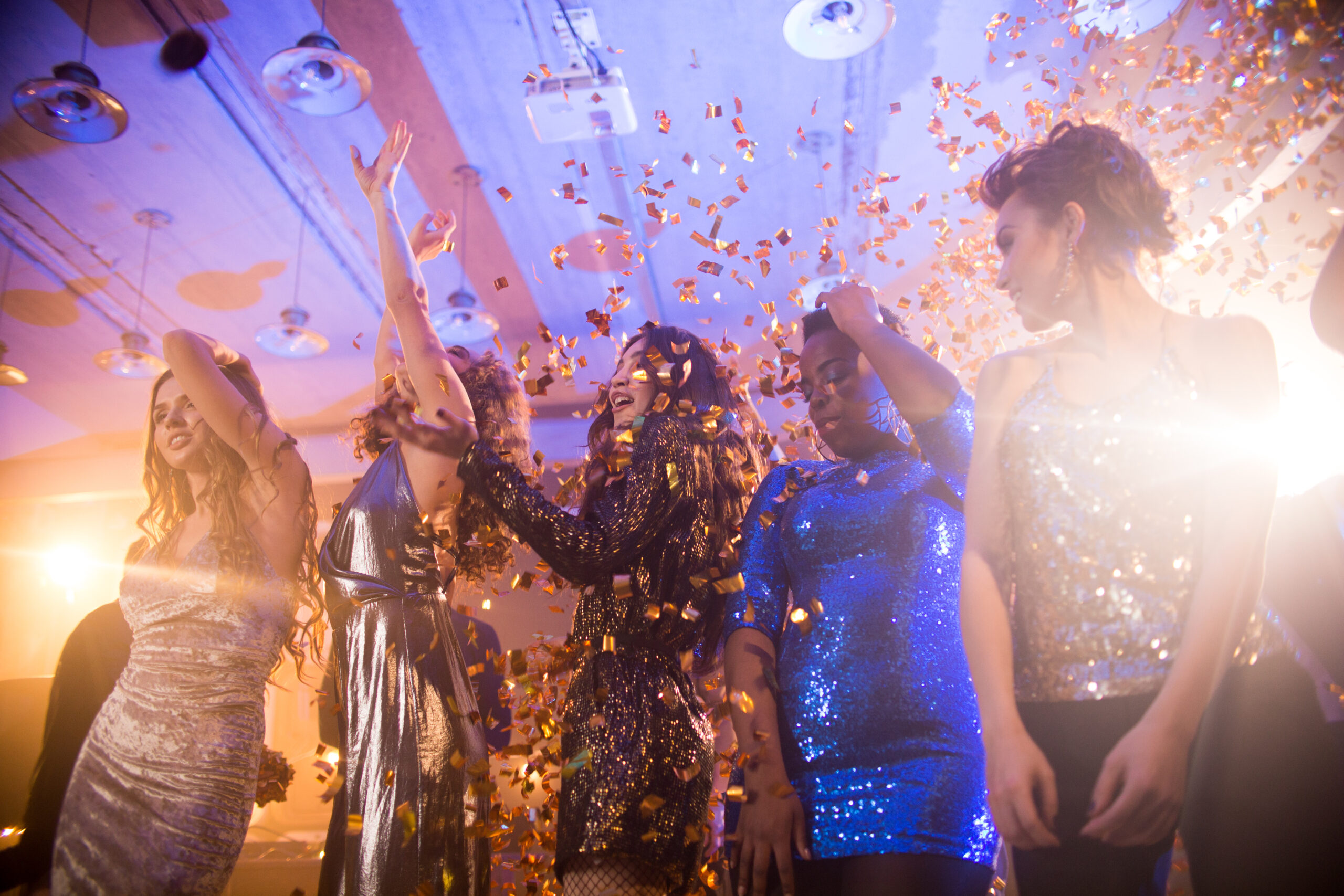 Group  of beautiful young women wearing glittering dresses dancing under golden confetti shower enjoying  party in nightclub